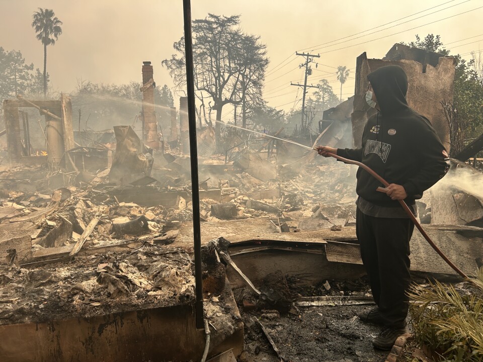 A person in a black hoodie and mask hoses down a burnt structure. 