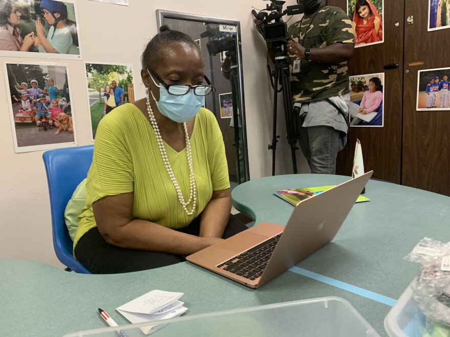 A woman in a green shirt and a blue paper mask conducts virtual teaching on a laptop.