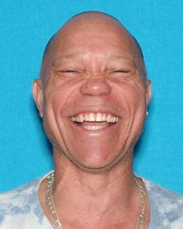A person with medium-tone skin and very short hair laughs in a mug shot.