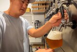 A man with medium in a light blue shirt and cap pours a golden beer from a tap into a tilted glass, creating a frothy head, with shelves of cans and equipment visible in the background.