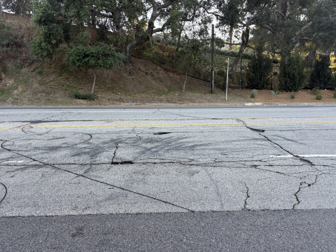 Cracks and patched potholes cover a paved road beside a tree-lined hillside.