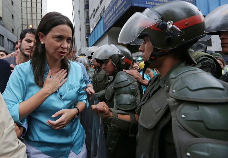 A woman with medium length brown hair, wearing a teal long sleeved shirt. She is holding her right hand to her heart as she speaks with a sodier wearing green protective gear. He is wearing a helmet with a red stripe and clear face shield attached to it