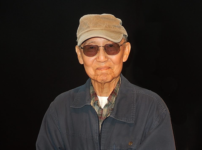 A Japanese American man wears sunglasses and a cap and is grinning. The backgound behind him is black.