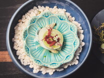Overhead view of an oyster dish in a turquoise, scalloped ceramic dish set in a blue bowl of white stones. A pale green disc is topped with red seaweed and a small white blossom.