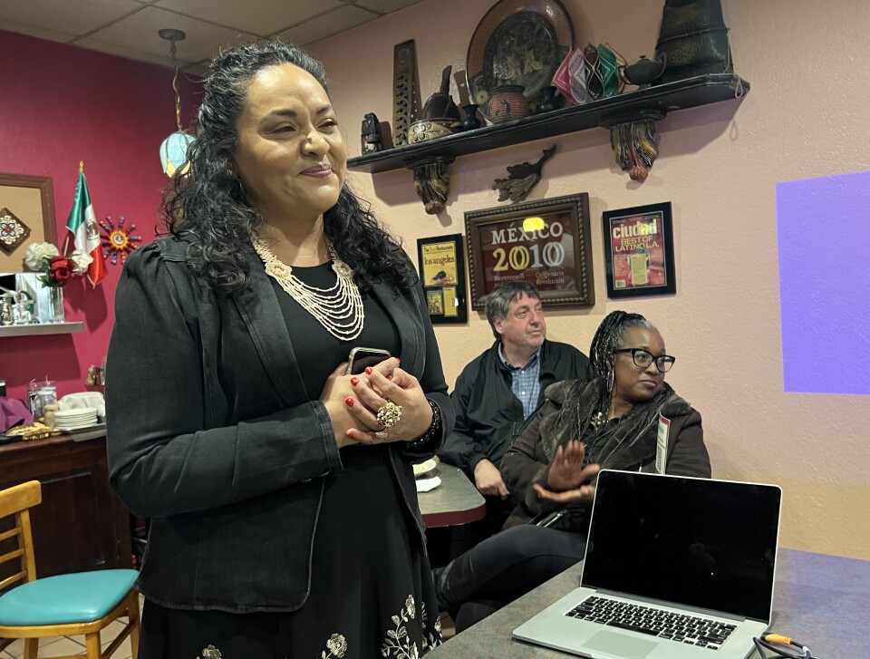 A woman with brown skin in a black dress wearing a light-colored necklace with roughly-shoulder-length brown hair smiles while an audience member applauds behind her.