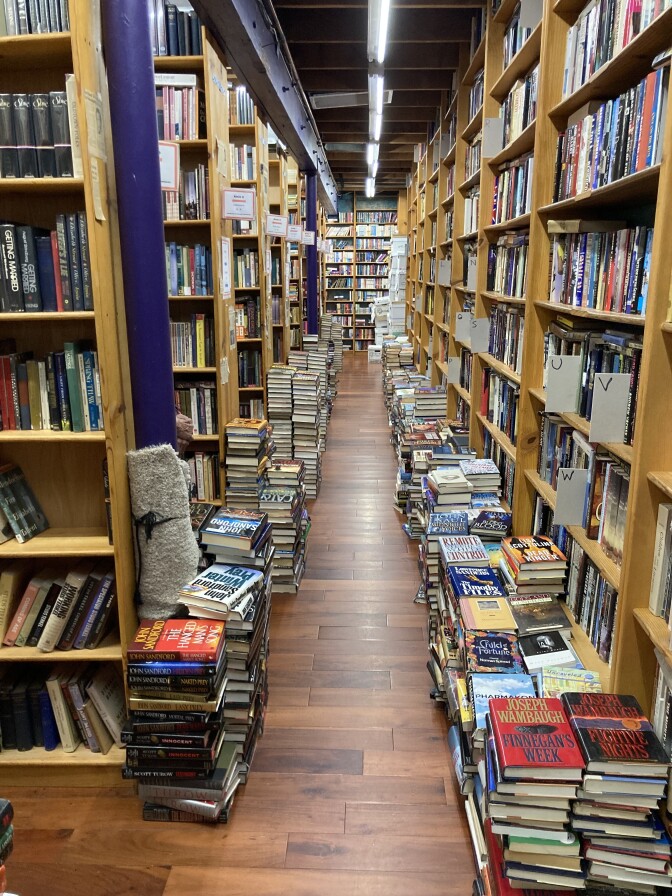 A view down a hallway of a used bookstore. There are rows of bookshelves on the left, a wall of shelves on the right, and stacks of books about 15-20 books high along either side of the hallway.