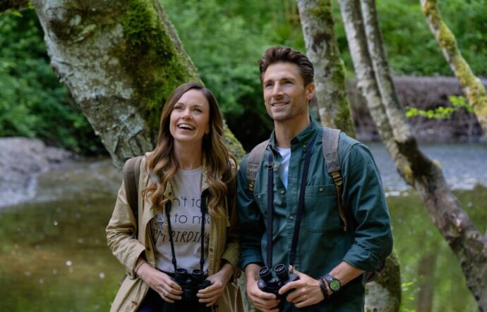 A man and woman stand together, each wearing backpacks and holding binoculars down at their waists. Behind them is a mossy tree trunk, grass and trees.