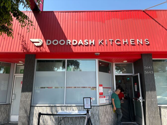 A building with a red roof with signage that says "DOORDASH KITCHENS" in white lettering. A man in a green shirt looks down at his phone and waits in the open doorway to the building. In front of the building, there's a white table accompanied by a sign that says: "ORDER CHECK TABLE ONLY."