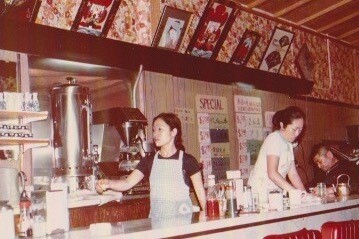 A grainy, vintage photo of two young Asian women behind a restaurant counter with red leather stools, serving customers.