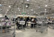 Fluorescent lights shine down on a busy floor where workers move trays on metal carts and sort ballots at a table