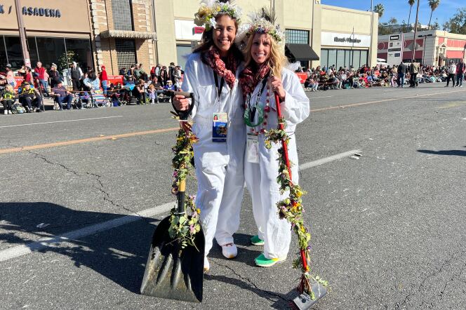 Two females, with light hair, wearing white jumpsuits and flower crowns, hold up a shovel and brush on a road. 
