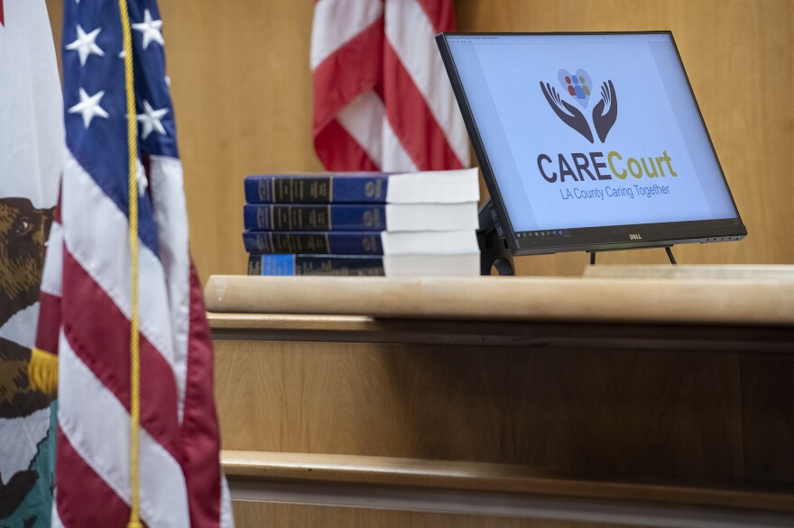 A computer screen inside a courtroom has graphics which read "CARE Court: LA County Caring together." An American flag and a California flag are to the left of the screen.