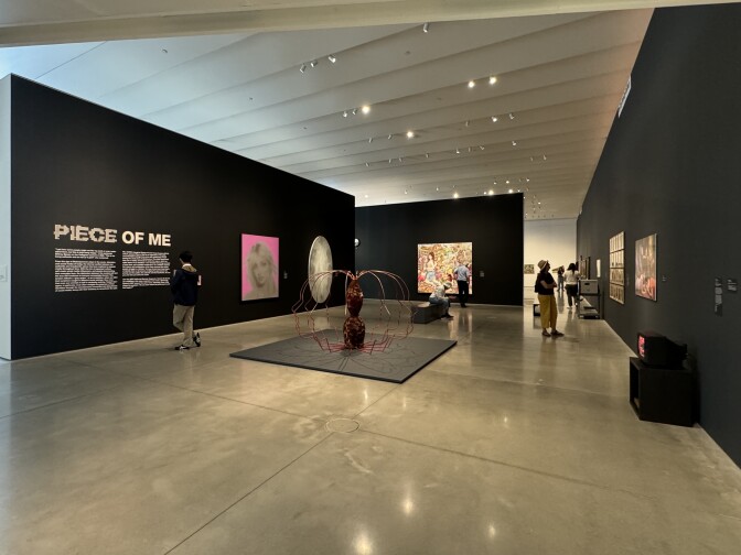 Scattered individuals look at black walls filled with different artwork. The title "Piece of Me" is displayed on the left wall. There is a black and white portrait of Britney Spears across a pink background among various other artworks.