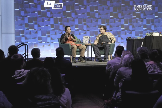 Chef Roy Choi who has a medium dark skin tone and LAist food writer Gab Chabrán who has a light skin tone and is wearing glasses speak to a packed audience at a Cookbook LIVE event. They're seated on stage with "The Choi of Cooking" book displayed between them. against a blue backdrop with LAist and James Beard Foundation branding.
