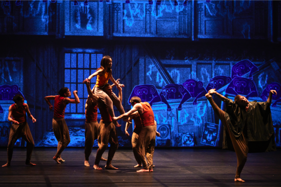 Seven dancers are on stage; one woman rests on the shoulders of another dancer. The rest of the dancers are caught in motion against a backdrop of blue illustration.