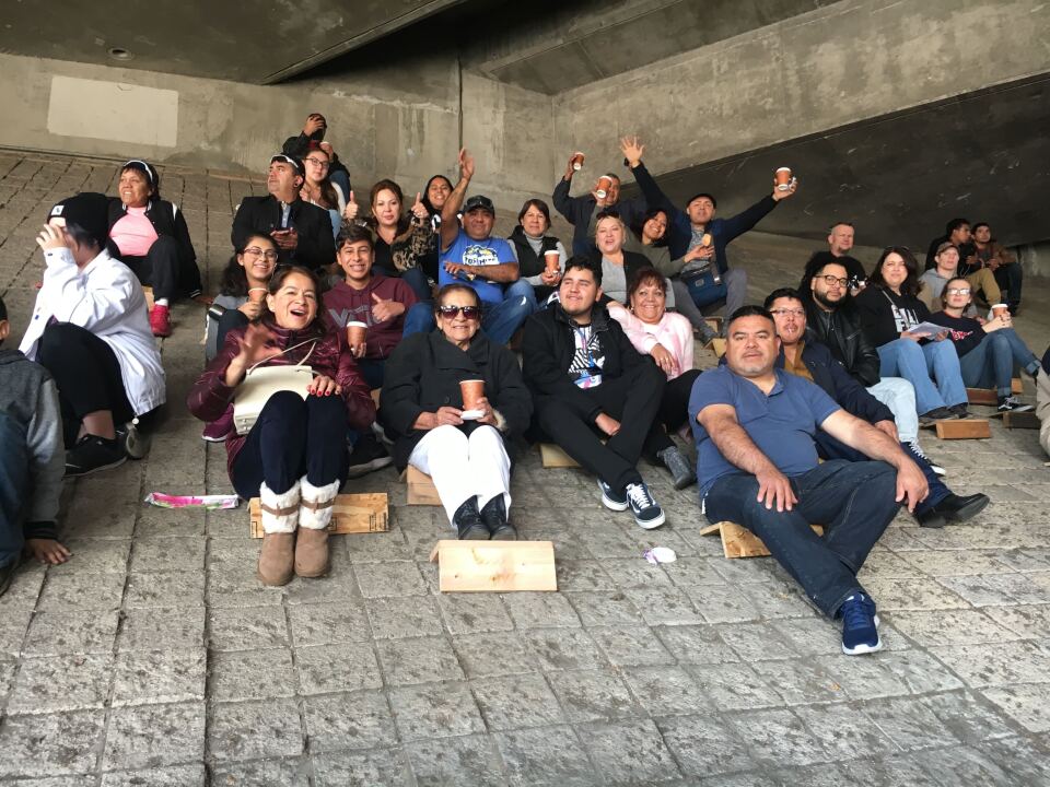 People sit on small wooden seats that help them perch on a sloped wall under a freeway overpass