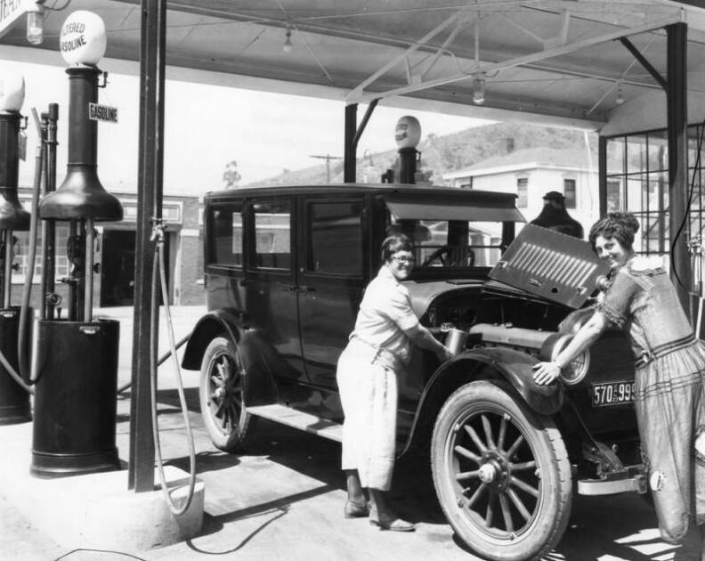 Two women in long dresses stand next to a car with its hood open.