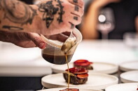 A pair of light skinned hands with several tattoos spoons a brown liquid over a series of white plates which each hold a piece of beef.