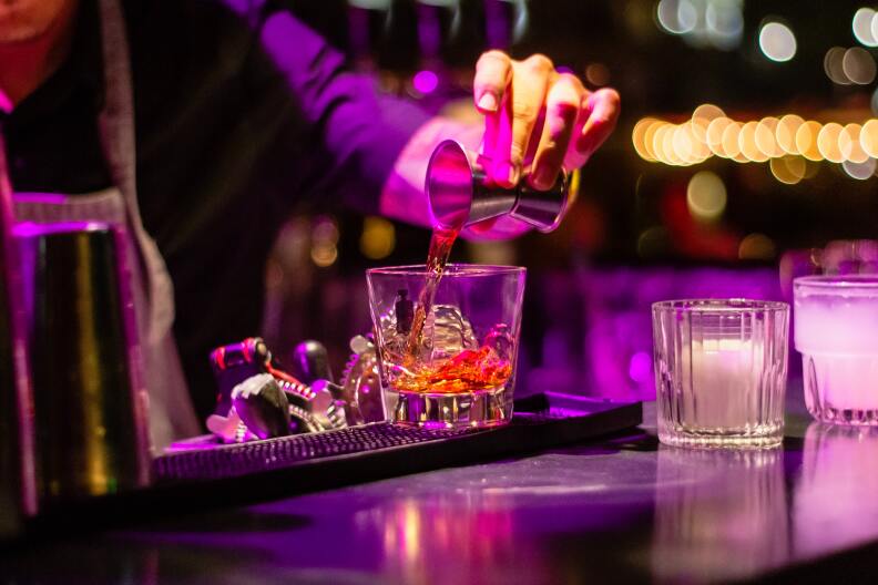 A bartender pours a shot into a cocktail glass. His arm is visible but not his face, and the scene is bathed in purple light.