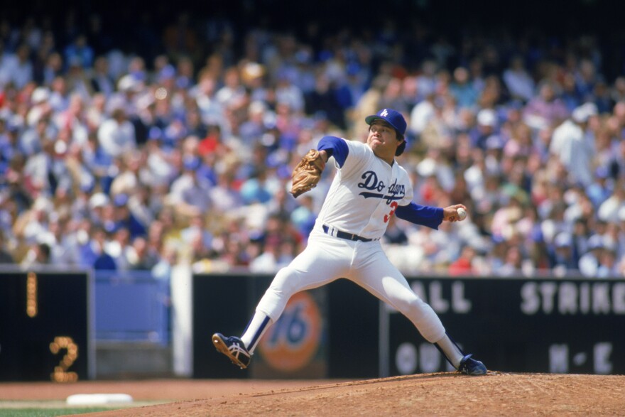 A pitcher in a white Dodger's uniform is mid wind-up on the pitcher's mound with a crowd in the stand behind.