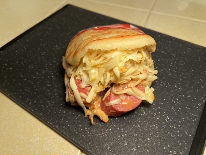 An image of an arepa, made with two white corn flour buns, containing a large pile of white grated cheese dressed with an orange sauce, with sliced pink sausage falling out. The arepa sits on a black-speckled cutting board on a yellow-tiled surface.  