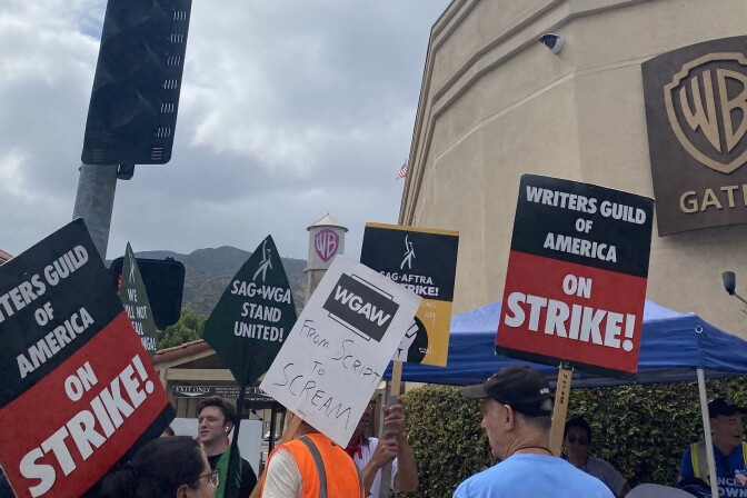 People holding signs that say "writers guild of america on strike" stand in front of a sign on the wall with the WB logo on it, Gate 4