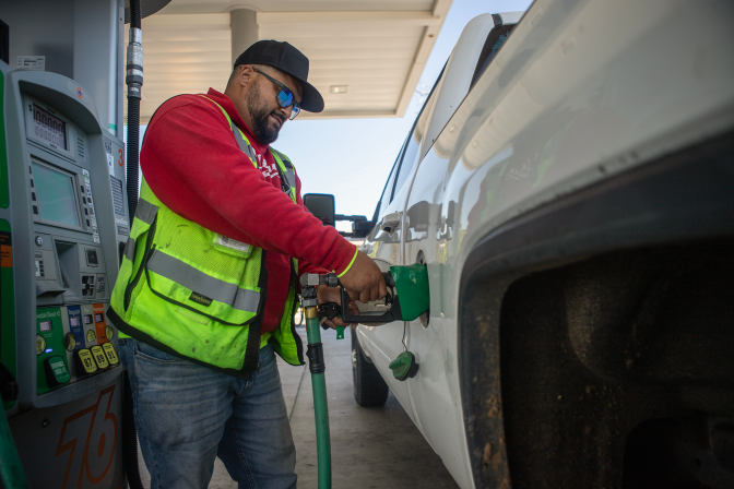 Man wearing read sweatshirt and fluorescent traffic vest pumps gas at gas station into a white truck