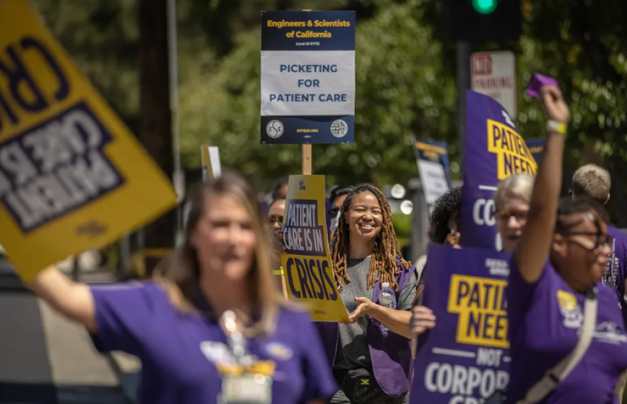 A group of protestors stand outside, holding various signs. In focus is a Black woman, near the back of the crowd. She has medium skin tone and is smiling at the camera. She has brown braids that are past her shoulder. She holds a yellow sign.