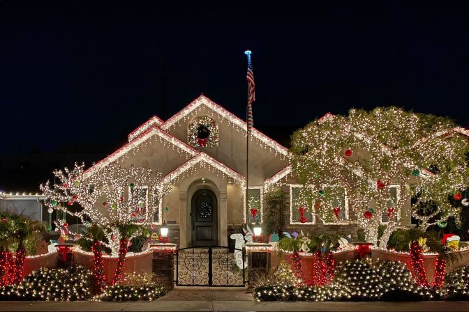 A home is decorated with white and red lights for the holidays. Two trees in front of the homes are also decorated with lights as are the bushes on the sidewalk. 