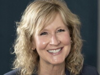 A woman with blonde, shoulder length hair, smiles while seated in front of a black background wearing a black blazer 