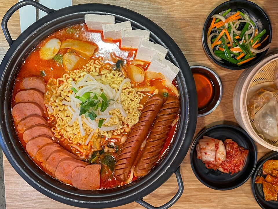 A large black pot and several smaller dishes are arranged on a light brown wooden tabletop. The large bowl is filled with a dark red liquid that contains a generous serving of curly yellow noodles at the center. It is topped with pieces of white and green onions, a curved row of sliced Spam, a row of white tofu, and two sliced hot dogs. To the right of the bowl, the smaller dishes include red kimchi cabbage, a light brown dipping sauce, and a small salad.