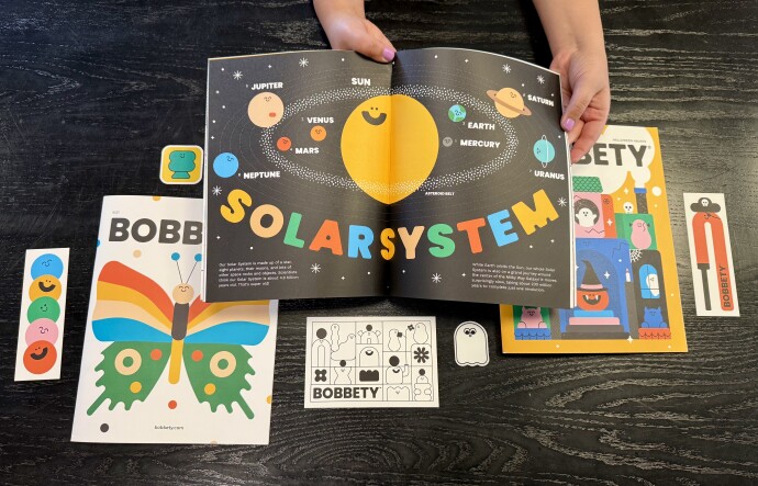 A pair of hands with medium light skin tone hold open a brightly colored magazine depicting the solar system with a bright yellow circle surrounded by smaller circles in different colors. The magazines in the background have a butterfly and ghosts on the cover. 