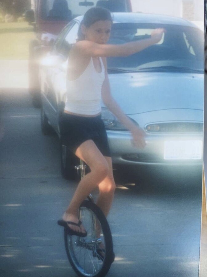 A young girl rides a unicycle in front of two parked cars.