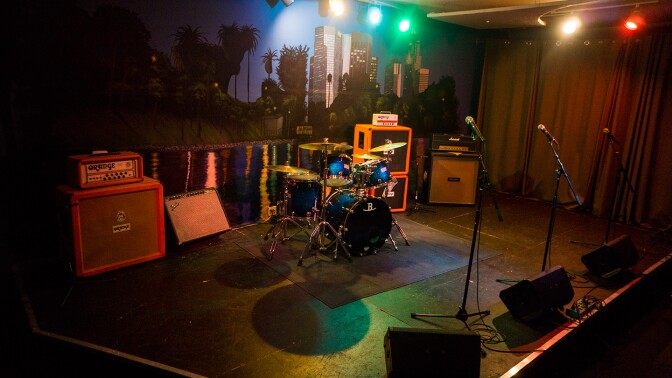 A small black stage with a curtain on one side, a cityscape backdrop on the wall. The stage has drums, amps, speaker monitors, and three microphones on stands.