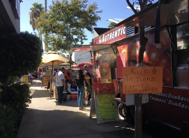 Signs, people, various stands and a red food truck line a city sidewalk.