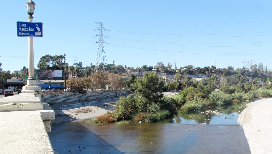View from the Fletcher Drive Bridge shows a blue sign with a heron reading "Los Angeles River" as well as foliage growing in the center of the river with graffiti-filled concrete banks beyond.