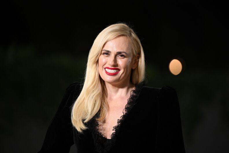 Actress Rebel Wilson smiles at the camera during an event. She's wearing black clothing and red lipstick.
