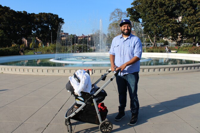A photo of a smiling man wearing a Dodgers hat, standing with a stroller in front of the fountain in the Rose Garden at Exposition Park