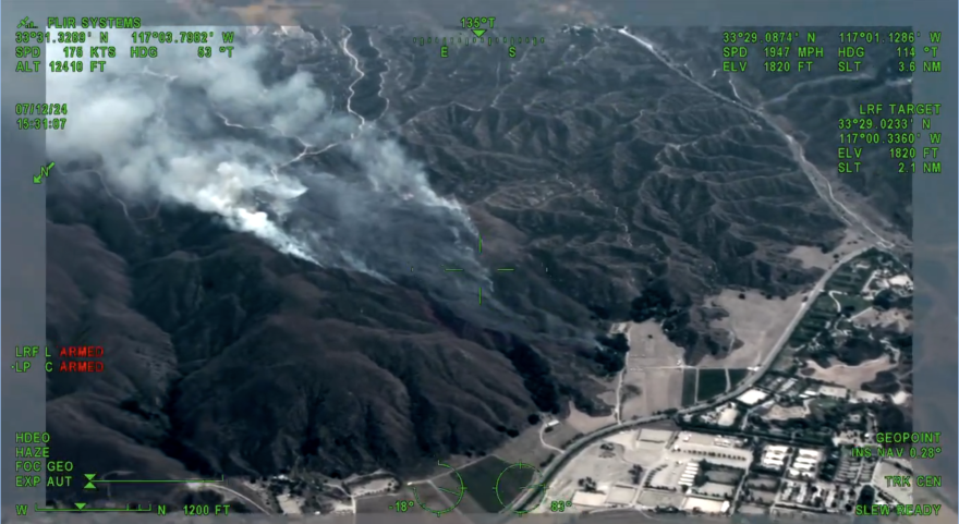 Smoke is seen rising inside the canyons of a rocky hillside. Buildings and roads can be seen at the foothills.