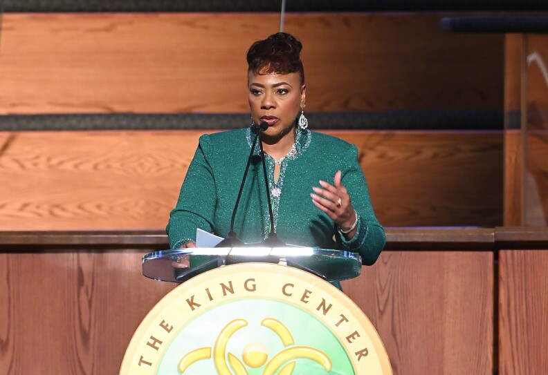 Bernice A. King stands at a lectern with the emblem "The King Center" She wears a green jewel-toned jacket and dangling earrings.