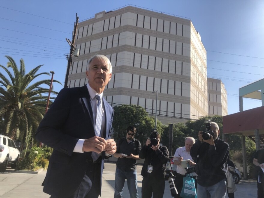 Wearing a blue suit and tie, George Gascón speaks to reporters when he first announced he was running for District Attorney. Reporters stand behind Gascón, holding cameras and writing in notepads. LA's Twin Towers Correctional Facility is in the background.