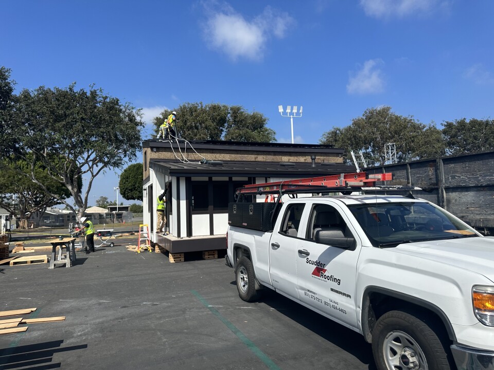 A 'Scudder Roofing' pickup truck sits at the foreground of the right side of the picture, behind it Rancho Cielo Construction Academy is underway putting the finishing touches on their submission to the Orange County Sustainability Decathlon. Two men work on installing the roof on the top of the home, one man is on the side of the home working on a window installation, on the asphalt below him another worker is cutting plywood for the installation. They are all wearing white hard hats and reflective yellow safety vests.