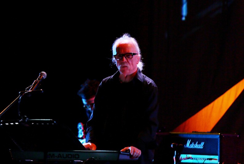John Carpenter, dressed in all black, is seen on stage with synths, a mic and a Marshall amp.