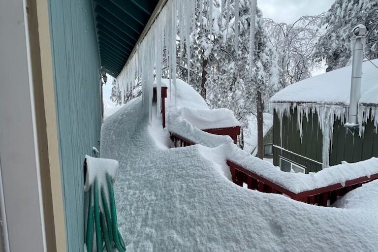 Snow covers the balcony and railing outside of a home. Snow lightly covers a garden hose hanging on the side of the house, with icicles coming down from the eaves above. White, snow covered trees are in the background, with more icicles and snow seen on another nearby neighboring home.