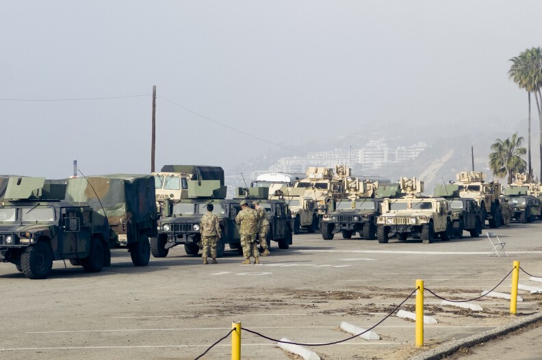 Three people in camo stand in front of dozens of armored vehicles in a parking lot. Pal trees and foggy hillside in the background.