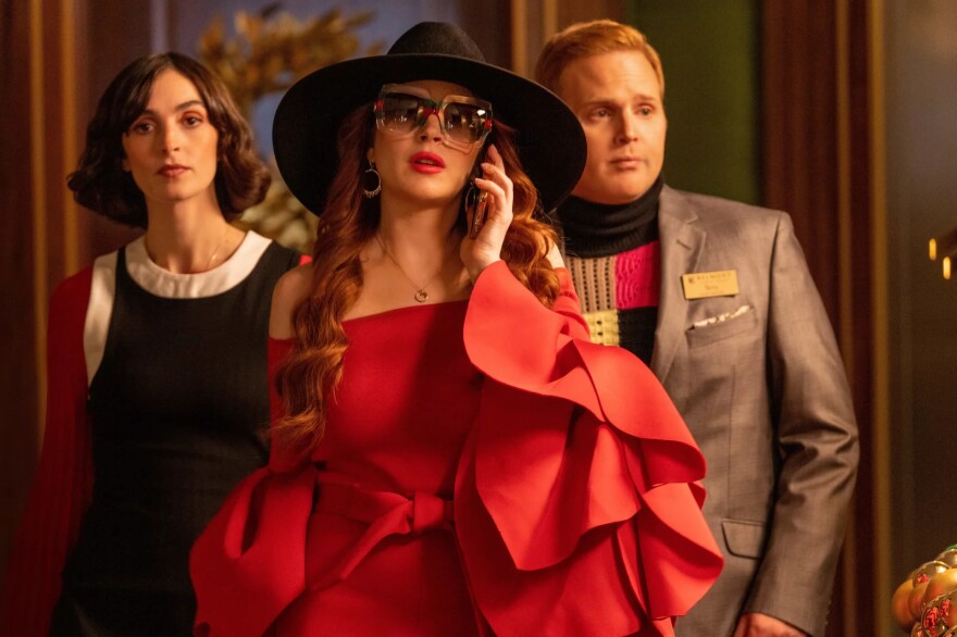 The actress Lindsay Lohan, a white woman who appears to be in her 20s or 30s, is shown in a movie still wearing an expensive-looking red dress, a large black hat. and sunglasses. She is speaking on a cell phone and trailed by a man and a woman.