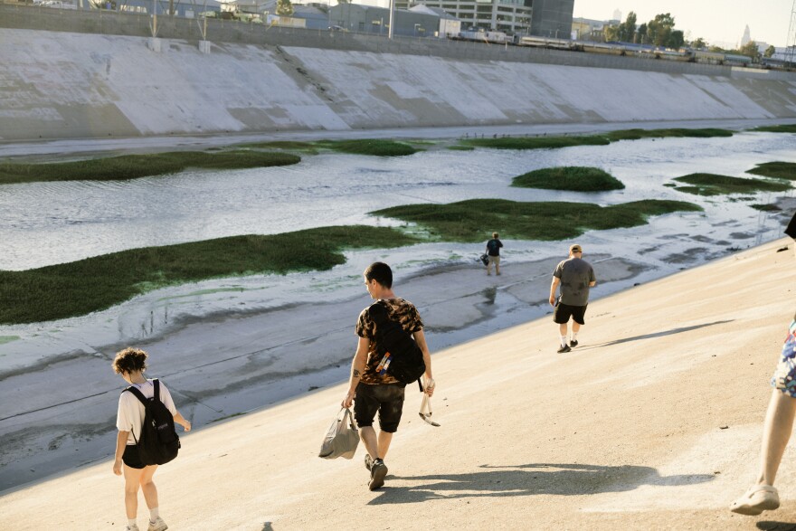 Multiple people approach the L.A. river cutting through a concrete structure. The river has some greenery and birds. A person in the center carries a backpack, bag, and tools.