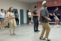 A dance studio with a handful of people spread out. At the front of the room is an instructor wearing glasses, a tan cap and a navy blue button-up shirt. 