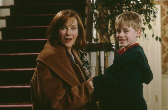 A woman wearing a brown coat and a young boy wearing a green hooded robe stand at the base of a stairwell inside a home.