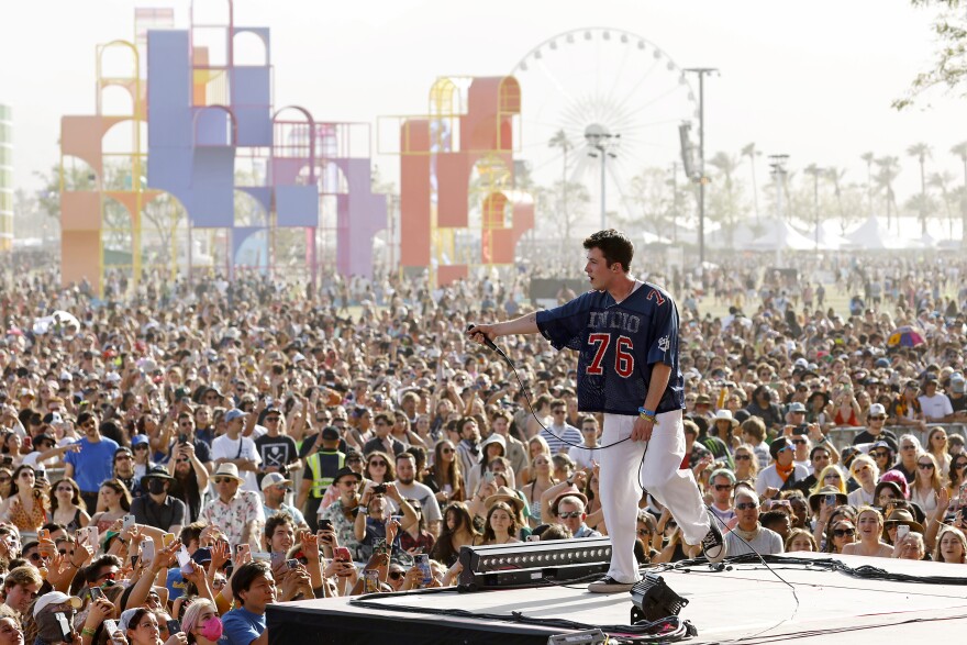 A young white man on a stage holds the microphone towards a massive outdoor crowd. He wears a mesh sports jersey with the number 76 and white pants. A colorful structure and a Ferris wheel can be seen in the background. It is daytime.